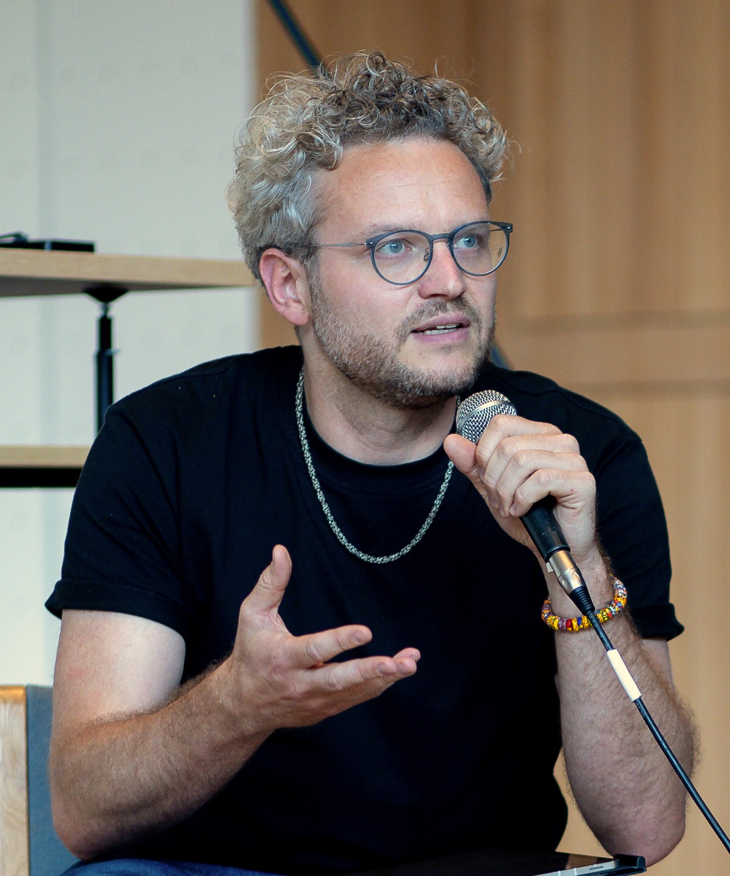 Leon Gabriel hält ein Mikrofon in der linken Hand und macht eine unterstützende Geste (nach oben geöffnete Hand) mit der rechten. Er schaut nach links und spricht. Dabei trägt er ein schwarzes T-SHirt, eine blaue Jeans, eine silberne Kette und eine blaue Brille.